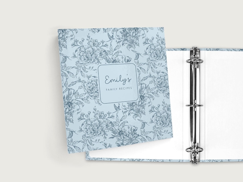 May include: A light blue binder with a floral pattern and the text "Emily's Family Recipes." The binder is open, revealing a three-ring mechanism and white pages. The floral design features flowers and leaves.