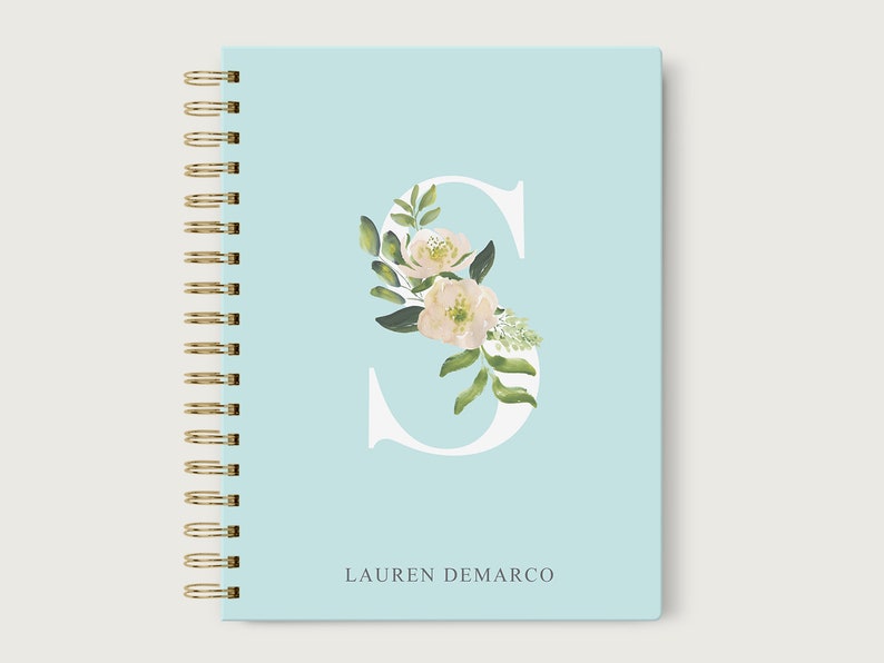 May include: A light blue spiral bound notebook with a white letter "S" decorated with white flowers and green leaves. The name "LAUREN DEMARCO" is printed in gray at the bottom of the notebook.
