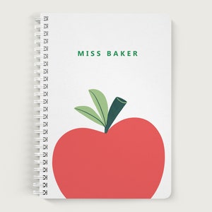 May include: A white spiral notebook with a red apple and green leaves on the cover. The text "MISS BAKER" is printed in green on the cover.