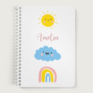 K&ouml;nnte beinhalten: Ein wei&szlig;es Spiralnotizbuch mit einer gelben Sonne, einer blauen Wolke und einem Regenbogen mit rosa, gelben und blauen Streifen. Die Sonne, die Wolke und der Regenbogen haben l&auml;chelnde Gesichter. Der Name Amelia ist in rosa Kursivschrift auf dem Notizbuch geschrieben.
