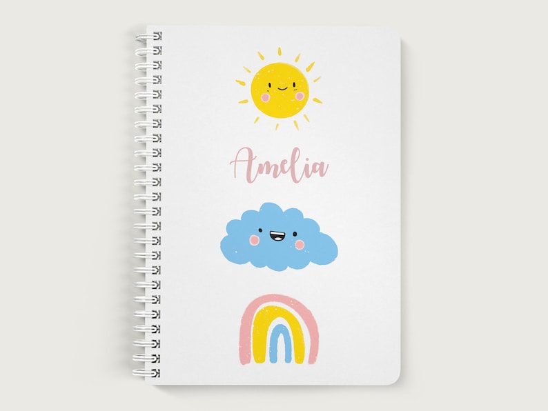 K&ouml;nnte beinhalten: Ein wei&szlig;es Spiralnotizbuch mit einer gelben Sonne, einer blauen Wolke und einem Regenbogen mit rosa, gelben und blauen Streifen. Die Sonne, die Wolke und der Regenbogen haben l&auml;chelnde Gesichter. Der Name Amelia ist in rosa Kursivschrift auf dem Notizbuch geschrieben.