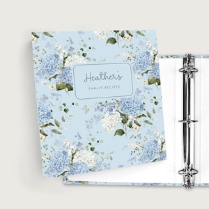 May include: A light blue three-ring binder featuring a floral pattern of blue and white hydrangea blossoms and green leaves. A central rectangular label on the cover displays the text Heather's FAMILY RECIPES in a dark blue script and sans-serif font.