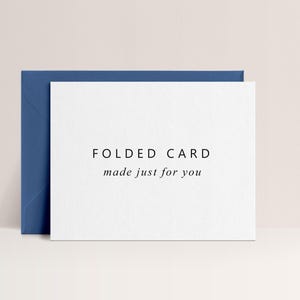 May include: A white folded card with the text "FOLDED CARD made just for you" in black font, resting against a dark blue envelope. The card and envelope are on a light beige background.