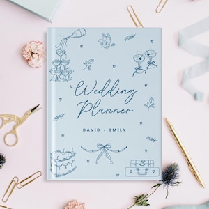 May include: A light blue wedding planner with the words "Wedding Planner" in elegant script. The cover features line drawings of champagne glasses, a cake, and luggage. The names "David + Emily" are printed below the title. Gold scissors, a pen, and paperclips are scattered around the planner.