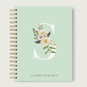 May include: A mint green spiral bound notebook with a white letter "S" decorated with white flowers and green leaves. The name "LAUREN DEMARCO" is printed in gray at the bottom of the notebook.