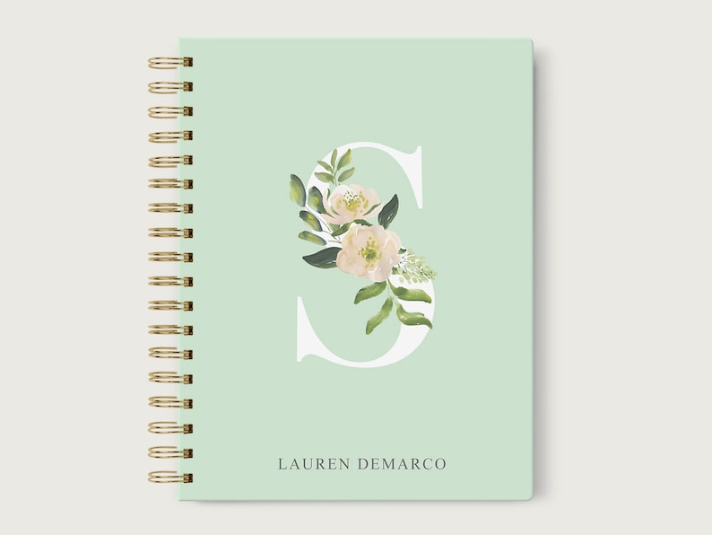 May include: A mint green spiral bound notebook with a white letter "S" decorated with white flowers and green leaves. The name "LAUREN DEMARCO" is printed in gray at the bottom of the notebook.