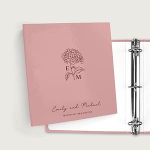 May include: A pink binder with a floral design and the initials "E" and "M". The text "Emily and Michael WEDDING ORGANIZER" is written in script below the floral design. The binder is open, revealing white pages.