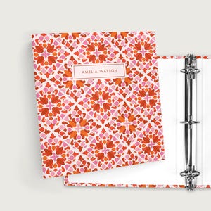 May include: A three-ring binder with a colorful floral pattern in shades of pink, orange, and white. The binder is open, revealing white interior pages and silver metal rings. The name "AMELIA WATSON" is printed on a label.