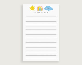 May include: A white notepad with a yellow sun, rainbow, and blue cloud with faces. The notepad has the name "Adeline Johnson" printed at the top.