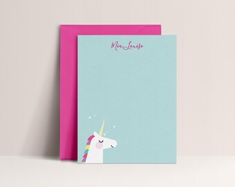 May include: A light blue notepad with a unicorn illustration and the name "Mia Louise" in pink script. A bright pink envelope is behind the notepad. The unicorn has a rainbow mane and a golden horn. The notepad has a few small white stars.