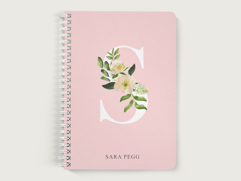 May include: A pink spiral bound notebook with a white letter "S" decorated with green leaves and white flowers. The name "SARA PEGG" is printed below the letter.