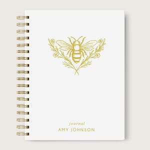 May include: White spiral-bound journal with a gold bee and floral design on the cover. The word "journal" and the name "AMY JOHNSON" are printed in gold below the bee illustration. The spiral binding is gold.