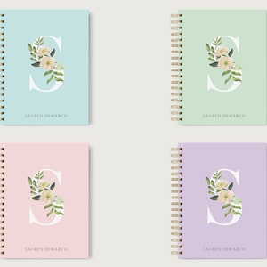 May include: Four spiral bound notebooks with different colored covers: light blue, light green, light pink, and light purple. Each cover features a white letter "S" with a floral design in shades of green and white. The name "LAUREN DEMARCO" is printed in black at the bottom of each cover.