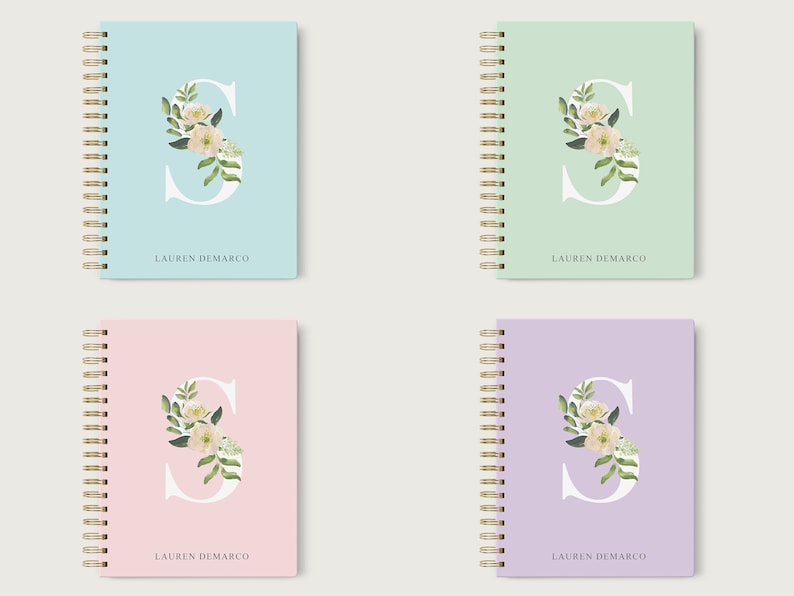May include: Four spiral bound notebooks with different colored covers: light blue, light green, light pink, and light purple. Each cover features a white letter "S" with a floral design in shades of green and white. The name "LAUREN DEMARCO" is printed in black at the bottom of each cover.