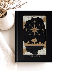 May include: A black journal featuring a tarot card design. The card depicts a gold star surrounded by smaller stars, clouds, and a landscape. The text "THE STAR" is at the bottom. The journal is on a white surface.
