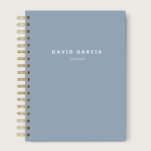 May include: A blue journal with gold spiral binding. The cover features the text "DAVID GARCIA" above "journal" in a minimalist font. The journal is closed and sits on a neutral background.