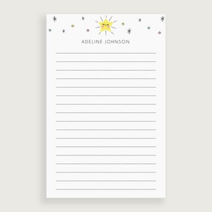 May include: A white lined notepad with a yellow star with a smiling face and a black outline. The star is surrounded by small colorful stars. The text "ADELINE JOHNSON" is printed at the top of the notepad.