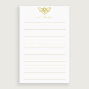 May include: A white notepad with gold lined paper and a gold bee illustration at the top. The name "AMY JOHNSON" is printed in gold below the illustration.