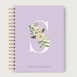 May include: A lavender notebook with a gold spiral binding. The front cover features a white letter "S" with a floral design in shades of green and white. The name "LAUREN DEMARCO" is printed in white at the bottom of the cover.