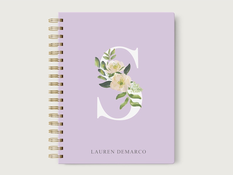 May include: A lavender notebook with a gold spiral binding. The front cover features a white letter "S" with a floral design in shades of green and white. The name "LAUREN DEMARCO" is printed in white at the bottom of the cover.