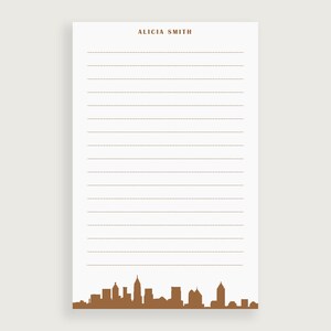 May include: A white lined notepad with the name "Alicia Smith" printed at the top. The bottom of the notepad features a brown silhouette of a city skyline.