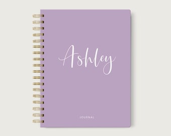 May include: A lavender spiral-bound journal with the name "Ashley" in white script. The word "JOURNAL" is printed at the bottom. The spiral binding is gold.