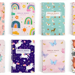 May include: Eight spiral-bound sketchbooks by Amelia Grace, each featuring a unique pattern including mermaids, rainbows, dogs, castles, fairies, unicorns, butterflies, and woodland animals, all with a white label displaying the brand name.