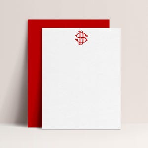 May include: White stationery with a red monogram and a red backing. The monogram is an ornate design in the upper center of the stationery. The stationery is leaning against a red background. The overall theme is stationery.