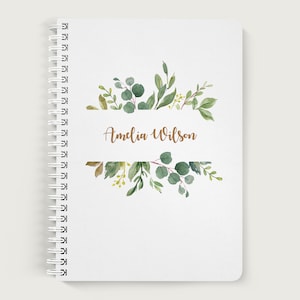 May include: A white spiral bound notebook with a copper colored name "Amelia Wilson" printed on a white banner surrounded by a watercolor design of green leaves.