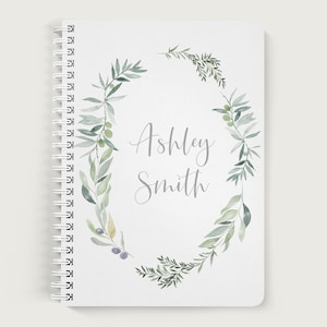 May include: A white spiral bound notebook with a watercolor wreath of green leaves and olive branches. The name "Ashley Smith" is written in gray script in the center of the wreath.