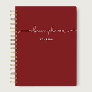 May include: A red journal with gold spiral binding. The cover features the name "rebecca johnson" in a cursive font, with the word "JOURNAL" below it in a sans-serif font. The journal is closed and sits on a neutral background.