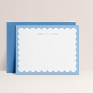 May include: A light blue stationery set featuring a scalloped edge card and matching envelope. The card has the name "AMELIA GRACE" printed in a light blue font. The stationery set is on a white surface.