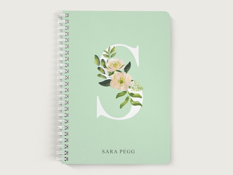 May include: A mint green spiral notebook with a white letter "S" decorated with watercolor flowers and leaves. The name "SARA PEGG" is printed in dark green at the bottom.