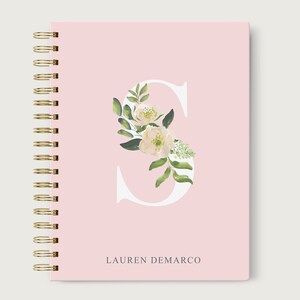 May include: A pink spiral bound notebook with a white letter "S" decorated with white flowers and green leaves. The name "LAUREN DEMARCO" is printed in gray at the bottom.