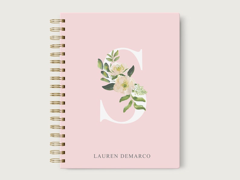 May include: A pink spiral bound notebook with a white letter "S" decorated with white flowers and green leaves. The name "LAUREN DEMARCO" is printed in gray at the bottom.