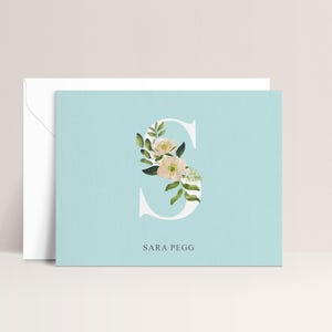 May include: A light blue stationery card with a white floral-filled letter "S" and the name "SARA PEGG" in a simple font. A white envelope is behind the card.