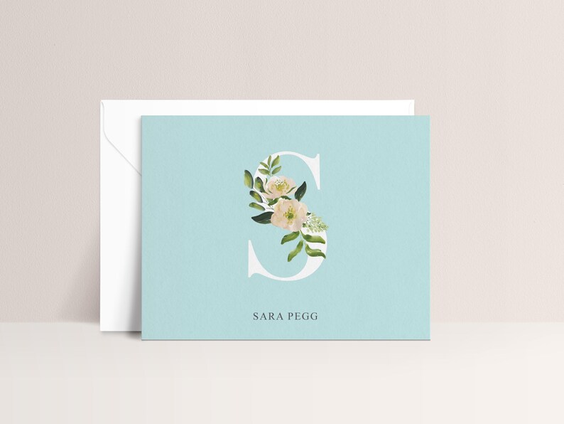 Puede incluir: Tarjeta de papeler&iacute;a azul claro con una letra "S" blanca llena de flores y el nombre "SARA PEGG". Se ve un sobre blanco detr&aacute;s de la tarjeta.