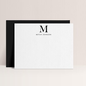 May include: A white stationery card with a black border, featuring the letter "M" and the name "Mitch Johnson" in a minimalist design. The card is set against a soft, neutral background, with a black envelope partially visible behind it. The overall aesthetic is clean and modern.