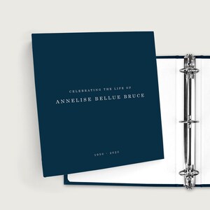 May include: A navy blue binder with silver metal rings, open to reveal white pages. The cover reads "Celebrating the Life of Annelise Bellue Bruce" with the years 1950-2025 in white text. A memorial or keepsake item.