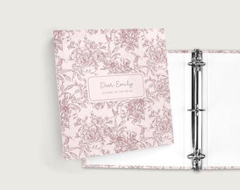 May include: A pink binder with a floral design and the text "Dear Emily Letters to the Bride." The binder is open, revealing a three-ring mechanism and white pages. The floral pattern is in a darker shade of pink.