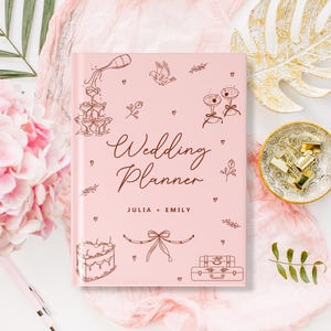 May include: A pink wedding planner with brown line art illustrations of champagne glasses, a cake, and luggage. The words "Wedding Planner" are written in a cursive font, with the names "JULIA + EMILY" below. The planner is surrounded by flowers and decorative items.