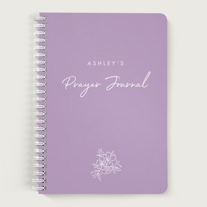 May include: A lavender-colored spiral bound journal with the text "ASHLEY'S Prayer Journal" in white lettering. A white floral design is at the bottom of the cover.