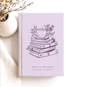 May include: A lavender reading journal with a stack of books and a teacup illustration. The cover features the text "AMELIA WILSON READING JOURNAL" in a serif font. A white vase with dried flowers is in the background.