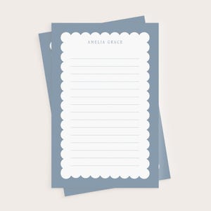 May include: Two blue notepads with a scalloped edge and lined paper. The top notepad has the name "AMELIA GRACE" printed at the top. The notepads are stacked on top of each other against a light beige background.