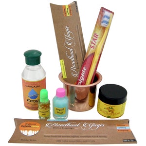 Puede incluir: Una colección de artículos de cuidado personal que incluye una botella de líquido transparente etiquetada como "GANGAJAL", una pequeña botella de aceite de sándalo, una botella de jabón líquido, un frasco de mascarilla facial de sándalo, una taza de cobre con un cepillo de dientes dentro y un paquete de inciensos etiquetados como "Avadhool Yogis Divine Blessings".