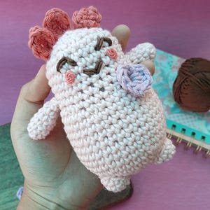 以下が含まれることがあります： 手作りの、薄いピンク色の編みぐるみのウーパールーパーのぬいぐるみ。茶色の刺繍の目と口、ピンクのアクセントがあります。ウーパールーパーは、側面に花があり、頭に装飾的なフリルが付いています。ぬいぐるみは手で持たれています。