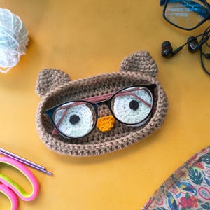 May include: A handmade, brown crochet owl-shaped glasses holder with black-framed glasses inside. The owl has white and black eye details and an orange beak. The holder sits on a yellow surface, with a ball of white yarn and scissors nearby.