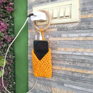 Può includere: Un portacellulare all'uncinetto giallo brillante con un telefono all'interno, appeso a una parete. Il telefono è collegato a un caricabatterie bianco. Il supporto è attaccato a un anello di legno che circonda un adattatore di alimentazione bianco.