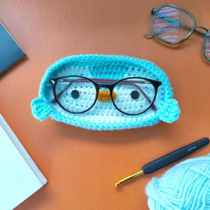 May include: A crocheted eyeglass holder in the shape of a penguin, with black-framed glasses inside. The holder is light blue with white and orange accents. A crochet hook and yarn are also visible.