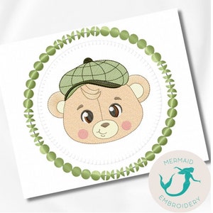 Puede incluir: Diseño bordado con un oso de dibujos animados con una gorra a cuadros verde, rodeado por un círculo de cuentas verdes. El oso tiene ojos marrones y mejillas rosadas. El diseño está sobre un fondo blanco, con un logo de sirena en la esquina inferior derecha.